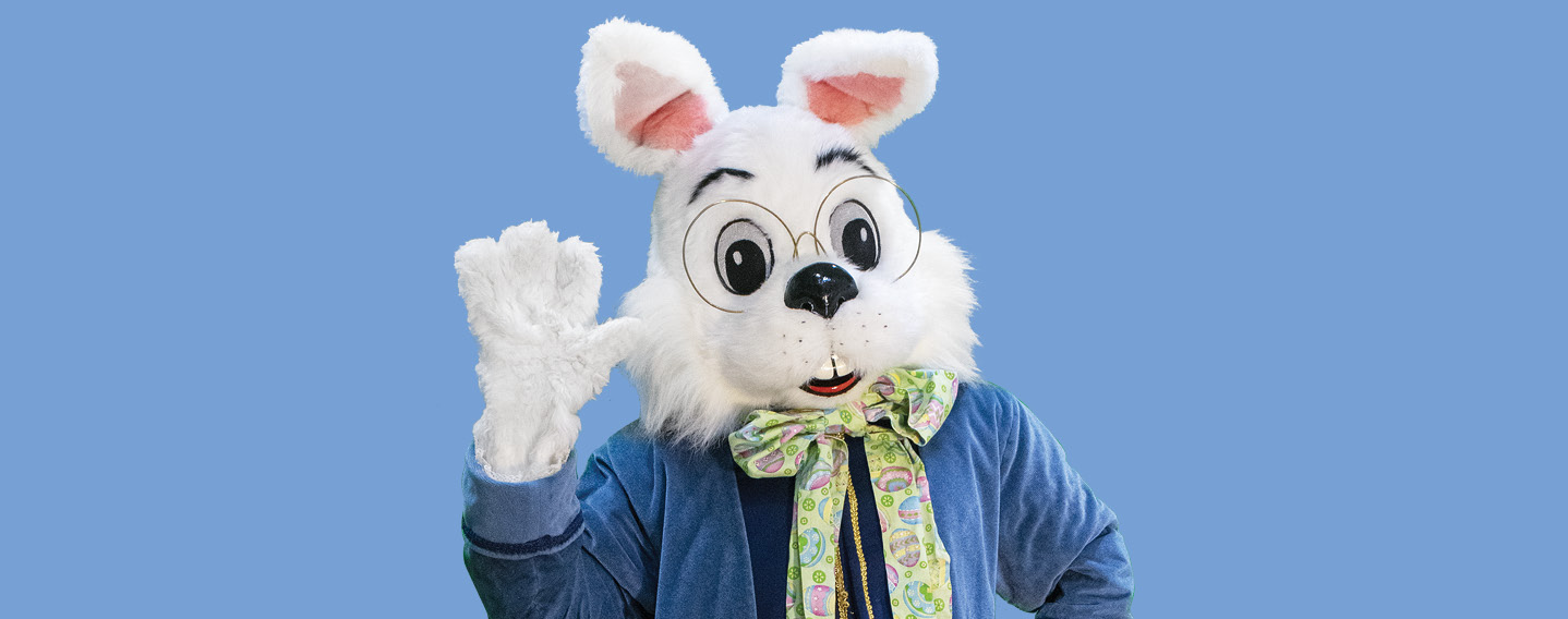 White bunny costume with glasses and a colorful bow tie waving for photos with Easter Bunny, set against a solid blue background.